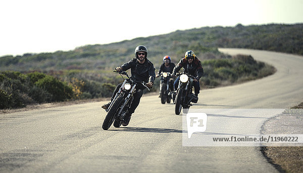 Three men wearing open face crash helmets and sunglasses riding cafe racer motorcycles along rural road.