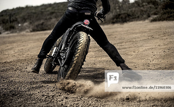 Rear view low section view of man riding cafe racer motorcycle on a dusty dirt road.