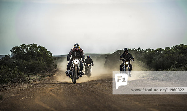 Three men riding cafe racer motorcycles along dusty dirt road.