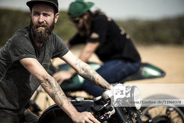 Side view of bearded man with tattoos on his arm sitting on cafe racer motorcycle on a dusty dirt road.