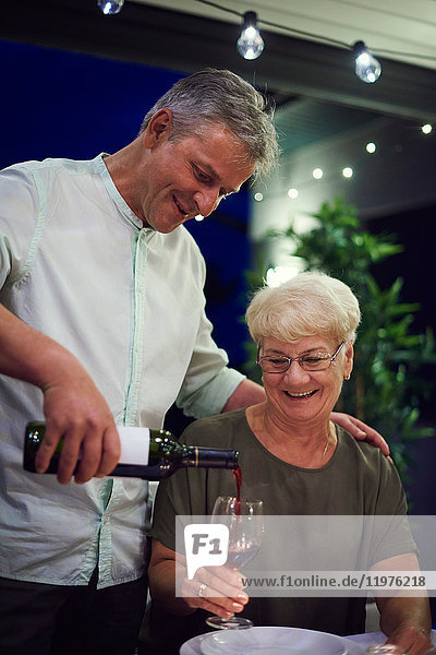 Ältere Frau sitzt am Esstisch  hält ein Weinglas in der Hand  reifer Mann gießt Wein ins Glas