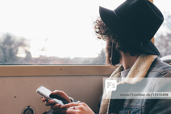 Junger Mann sitzt in der U-Bahn  hält ein Smartphone in der Hand und schaut aus dem Fenster