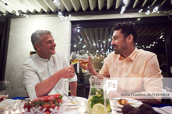 Two men sitting at dinner table,  holding wine glasses,  making toast
