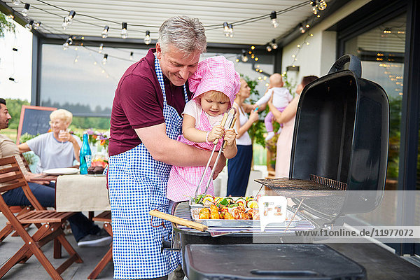 Female toddler barbecuing with grandfather at family lunch on patio