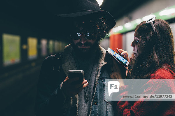 Junges Paar sitzt auf dem U-Bahnsteig und schaut auf Smartphones