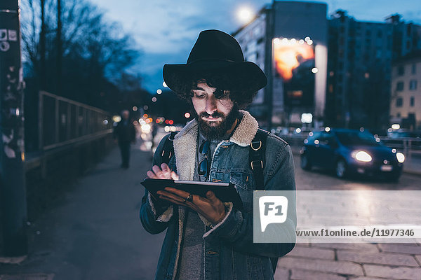 Junger Mann steht in der Abenddämmerung auf der Straße und benutzt ein digitales Tablett