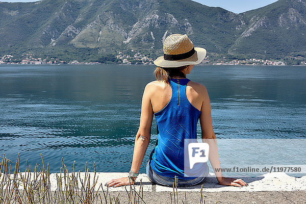 Rückansicht einer am Wasser sitzenden Frau in Trilby mit Blick auf die Bucht von Kotor  Montenegro