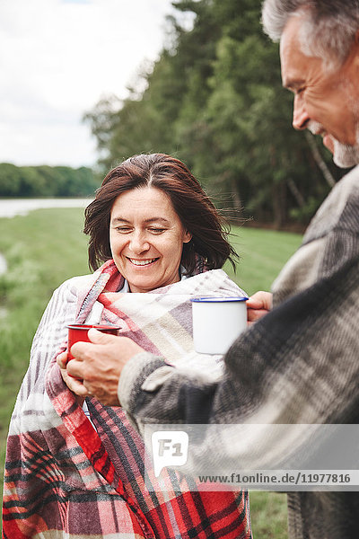 Reifes Paar steht in ländlicher Umgebung  hält heißes Getränk in der Hand  Frau in Decke gewickelt