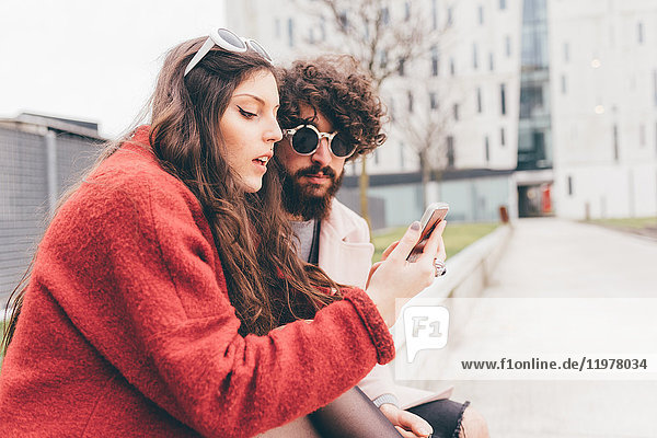 Young couple sitting outdoors looking at smartphone