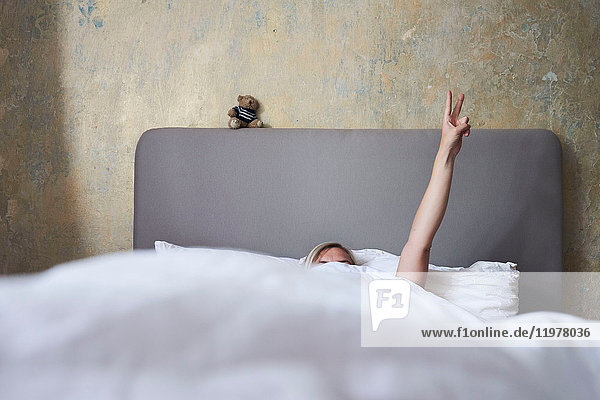 Frau im Bett  versteckt sich unter der Decke  Arm in der Luft  Hand zeigt Friedenszeichen