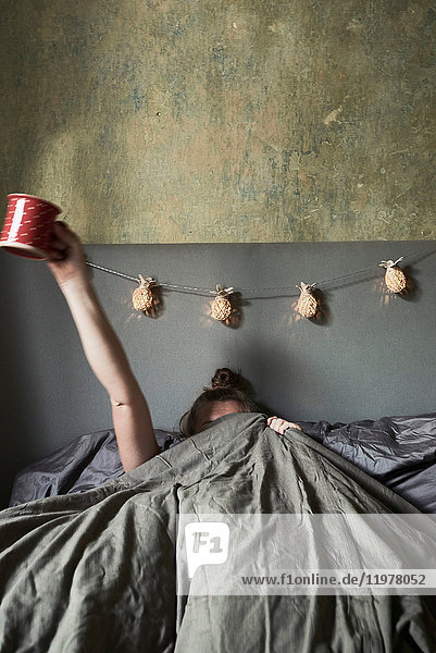 Woman in bed  hiding under covers  holding mug in air