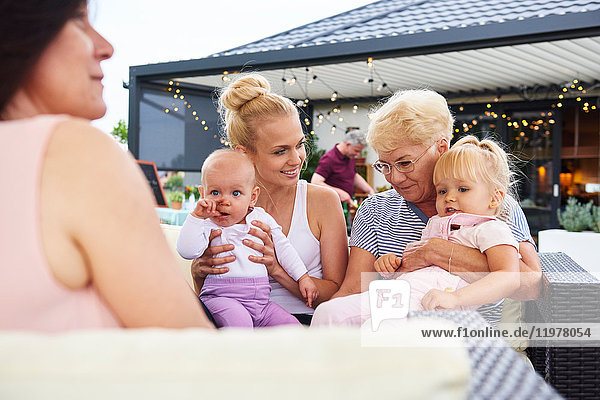 Senior and young women with female toddler and baby girl on laps at family lunch on patio