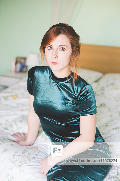 Portrait of young woman in shiny green dress sitting on bed