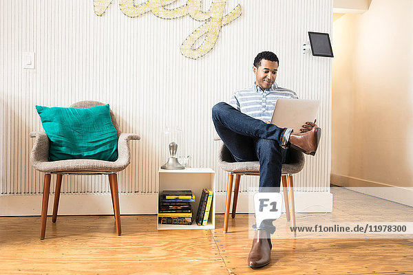 Cool young businessman sitting in creative office armchair using laptop
