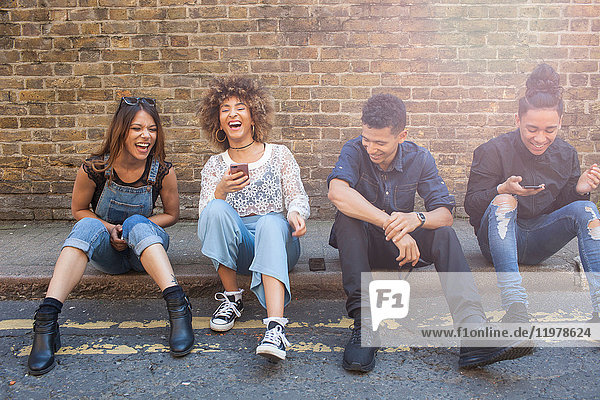 Four friends sitting in street  laughing  young woman holding smartphone