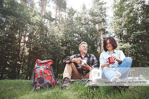 Mature couple relaxing on grass  holding tin cups  rucksack beside them