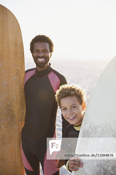 Portrait lächelnder Vater und Sohn mit Surfbrettern am sonnigen Sommerstrand