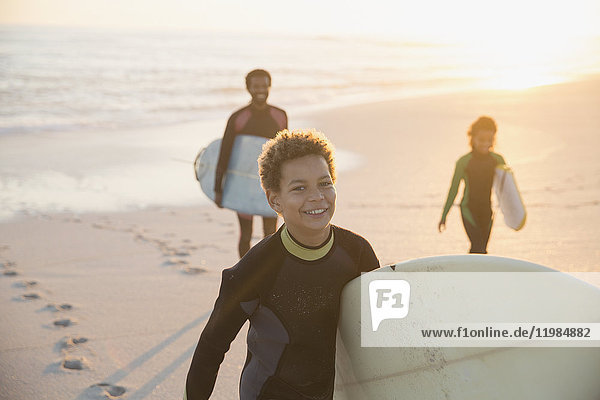 Portrait smiling pre-adolescent boy in wet suit carrying surfboard on summer sunset beach with family
