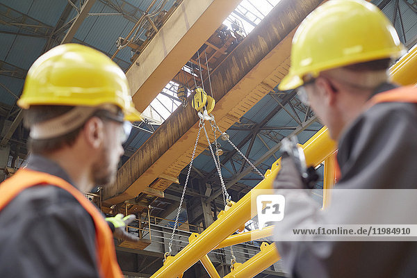 Male worker using walkie-talkie to guide hydraulic crane lowering equipment in factory