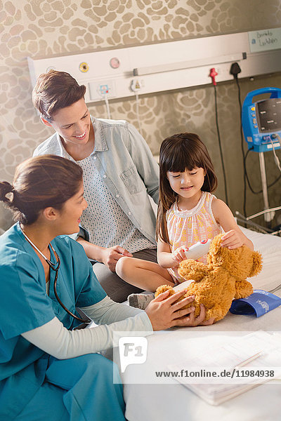 Eine Krankenschwester und ein Mädchen benutzen ein digitales Thermometer an einem Teddybären im Krankenhauszimmer