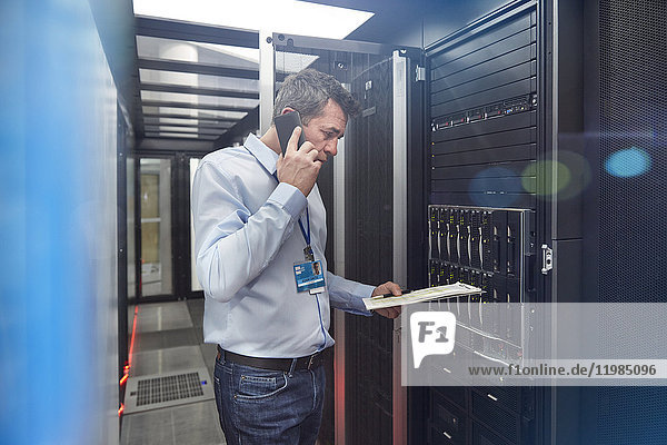 Male IT technician with clipboard talking on cell phone at panel in server room