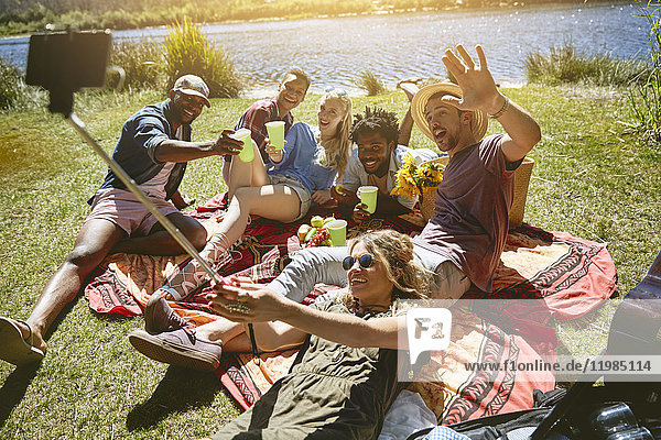 Verspielte junge Freunde nehmen Selfie mit Selfie-Stick  genießen Picknick am sonnigen Sommerufer