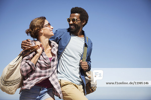 Affectionate multi-ethnic couple hugging and walking below sunny summer blue sky