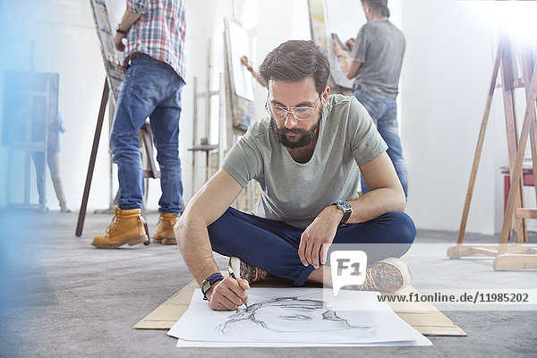 Male artist sitting cross-legged sketching on floor in art class studio