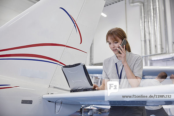 Flugzeugingenieurin bei der Arbeit am Laptop und beim Telefonieren im Hangar