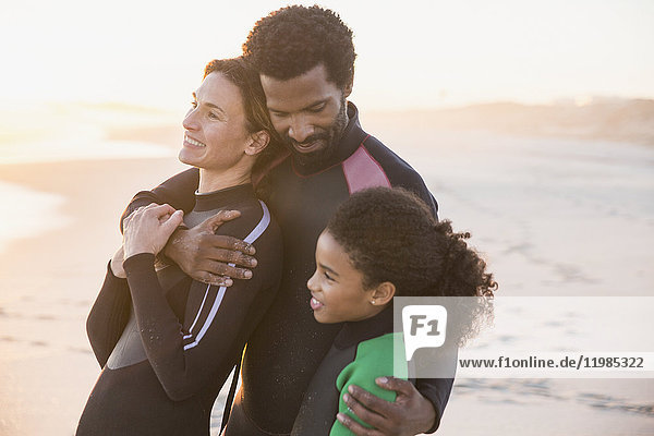 Liebevolle multiethnische Familie in Neoprenanzügen am Strand bei Sonnenuntergang