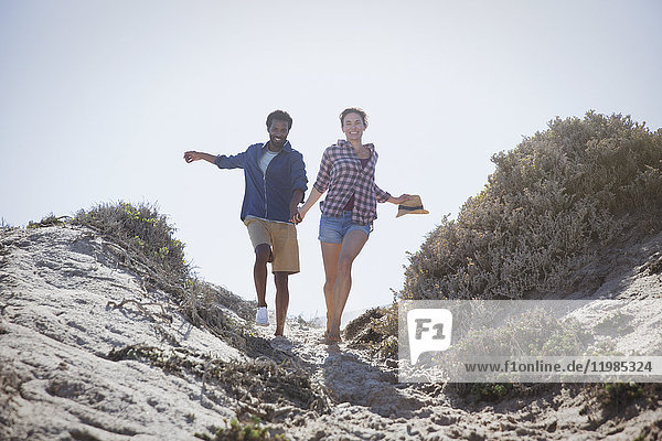 Verspieltes  energiegeladenes  multiethnisches Paar  das auf einem sonnigen Sommerstrandweg läuft und Händchen hält.