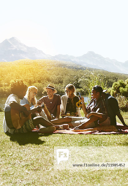 Junge Freunde hängen herum,  spielen Gitarre und genießen ein Picknick im sonnigen Sommergras.