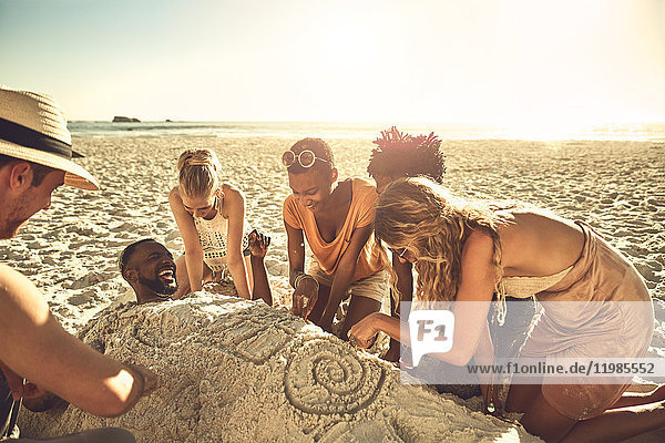 Young friends burying and drawing friend in sand on sunny summer beach