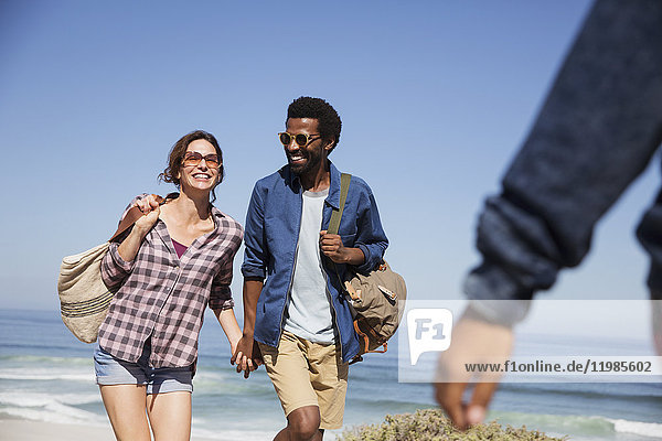 Lächelndes multiethnisches Paar  das Händchen hält und am sonnigen Sommerstrand des Ozeans spazieren geht.