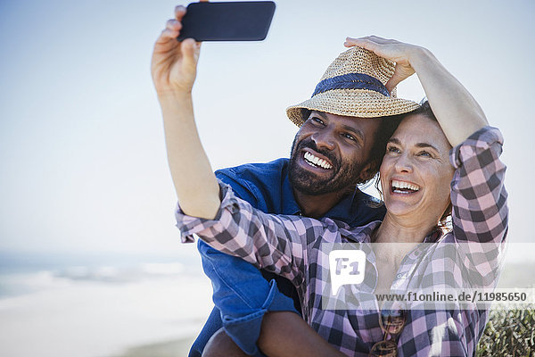 Verspieltes  lächelndes multiethnisches Paar  das sich selbst mit Fotohandy am sonnigen Sommerstrand fotografiert.