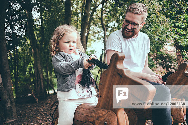 Happy father looking at daughter riding wooden horse in park
