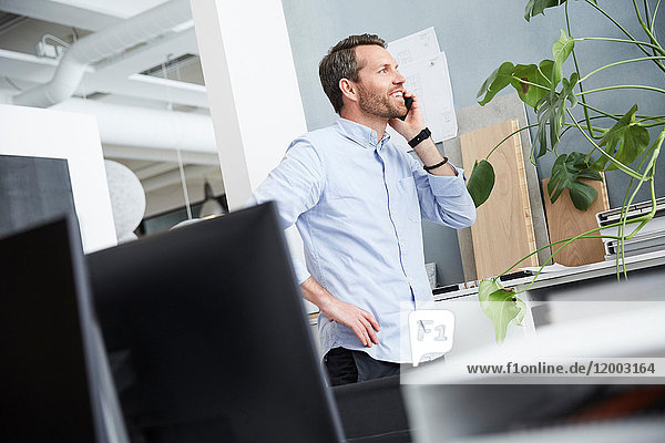 Smiling businessman talking on mobile phone while standing with hand on hip at office