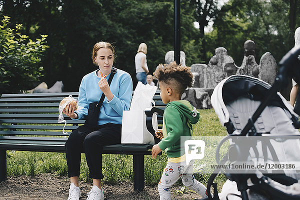 Mutter sitzt auf der Bank und isst  während sie den Sohn im Park sieht.
