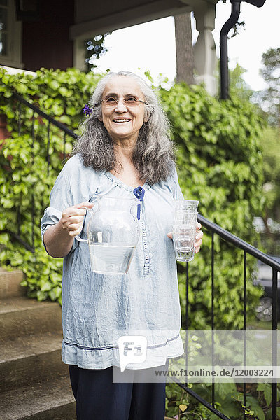 Lächelnde Seniorin stehend mit Wasserkrug und Glas im Garten