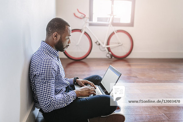 Man using laptop sitting on wooden floor with bicycle in background