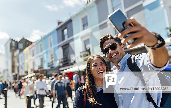 UK  London  Portobello Road  Porträt eines lachenden Paares  das sich selbst mit dem Smartphone fotografiert.