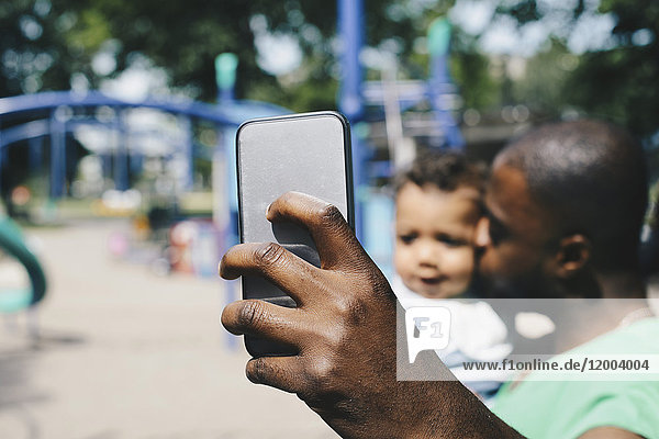 Father taking selfie through smart phone while kissing son at park