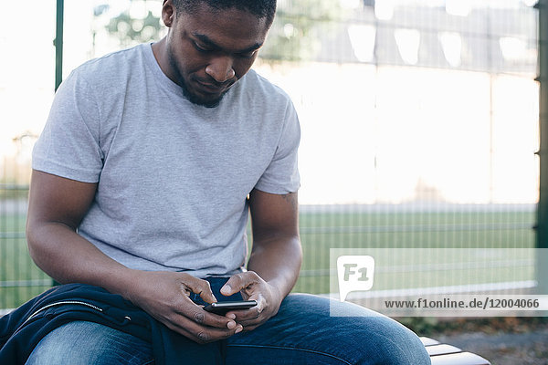 Junger Mann mit Smartphone auf der Bank gegen den Zaun sitzend
