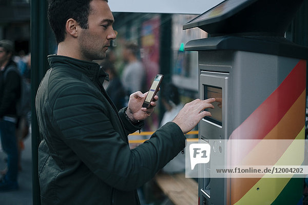 Seitenansicht des Mannes  der den ATM-Bildschirm berührt  während er das Smartphone hält.