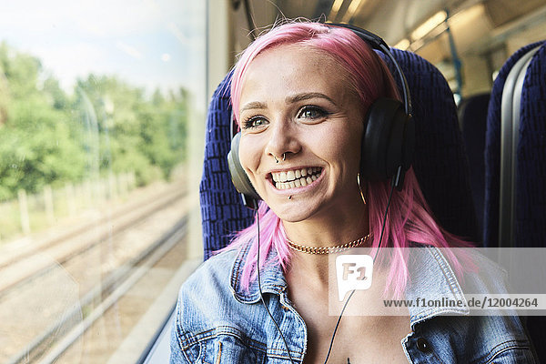 Fröhliche junge Frau mit rosa Haaren  die Musik hört  während sie mit dem Zug unterwegs ist.