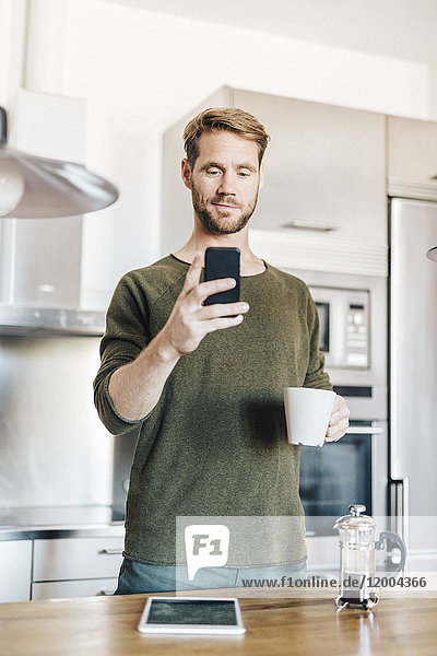 Porträt eines in der Küche stehenden Mannes mit Tasse Kaffee und Selbstbedienung mit Smartphone