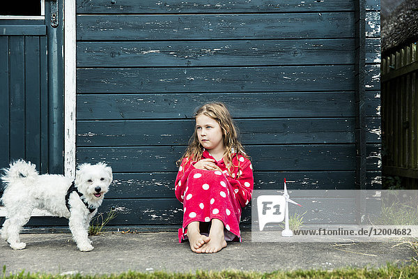 Mädchen im Bademantel im Freien sitzend an einer Holzhütte mit Spielzeug-Windkraftanlage und Hund