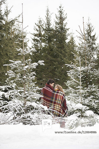 Glückliches junges Paar in Decke gehüllt im Winterwald stehend