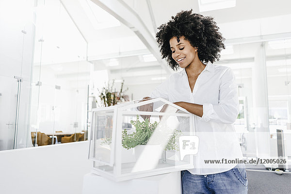 Smiling young woman in office caring for plants in glass box