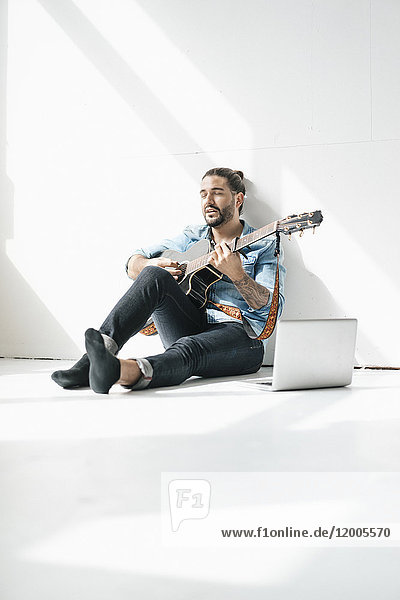 Singender Mann sitzend mit Laptop auf dem Boden und Gitarre spielend